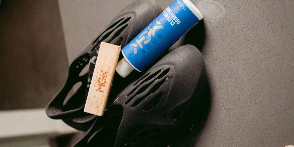 A bottle of the Shoe MGK cleaner and Conditioner leaning against pair of Yeezy Foam Runners