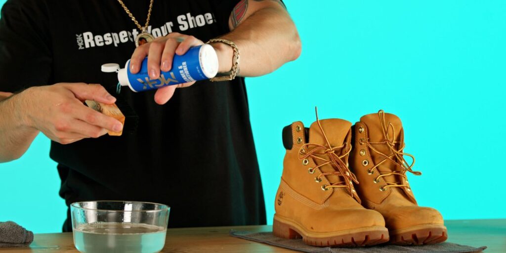 Man pours Shoe MGK Cleaner and Conditioner onto a nylon shoe brush above a bowl of water next to a pair of Timberland boots