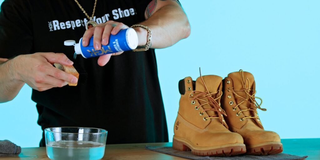 Man pours the Shoe MGK Cleaner and Conditioner onto a nylon brush over a bowl of water next to Timberland boots