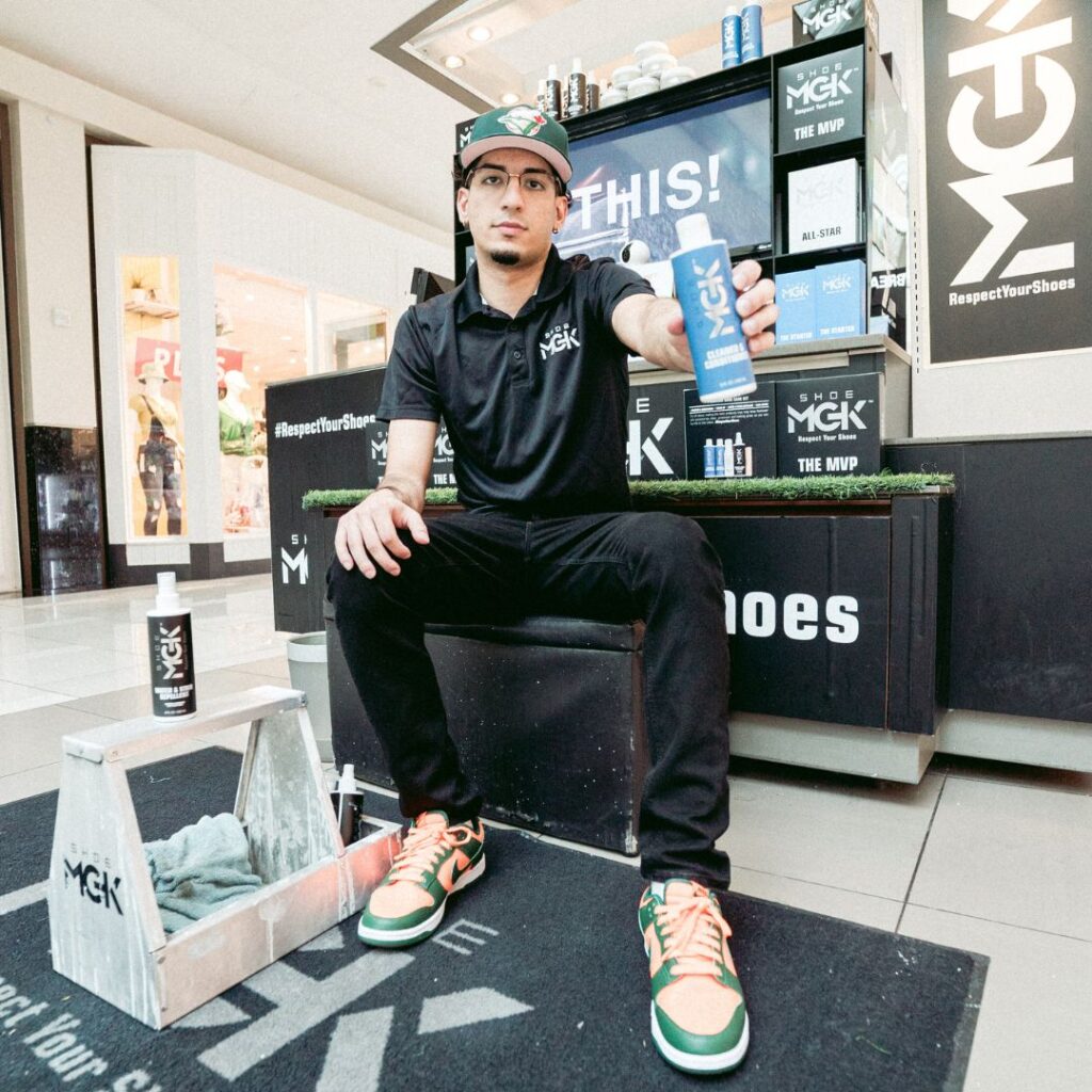 Man holds out a bottle of the Shoe MGK Cleaner and Conditioner while sitting at a mall kiosk